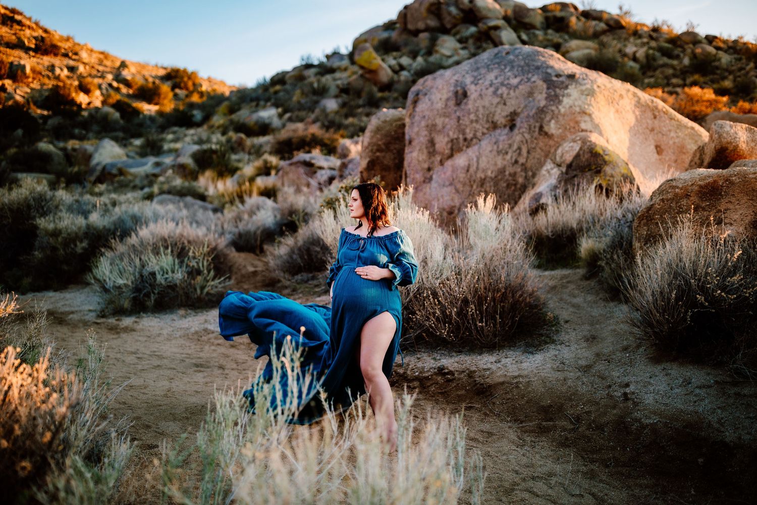 Maternity photo in a blue dress against desert rock formations at golden hour.