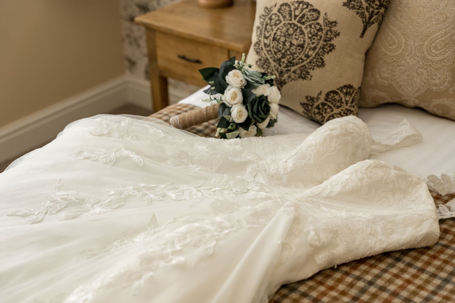 An elegant white lace wedding dress laid out on a bed with a matching floral bouquet.