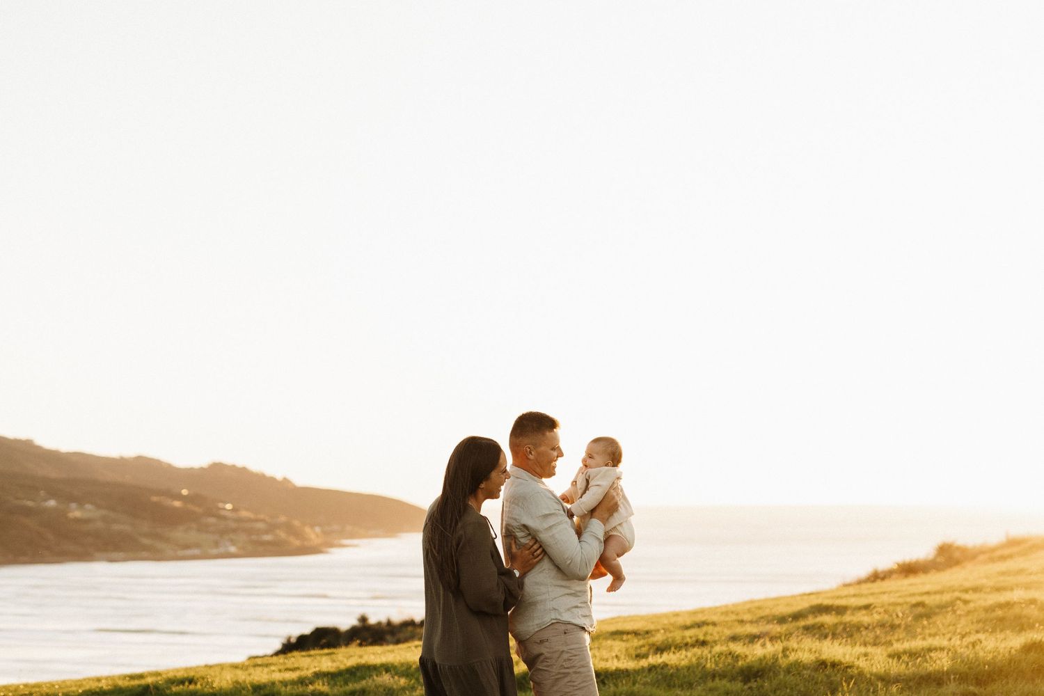 A family standing together on a grassy hill overlooking the ocean at sunset.