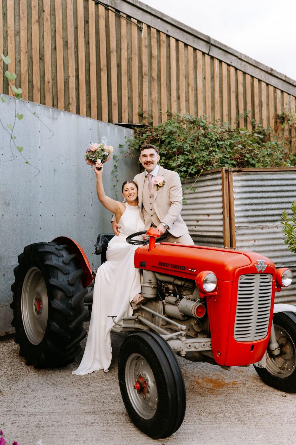 Bride and groom sitting on a red vintage tractor, bride holding bouquet aloft, groom in tan suit, at rustic barn venue.