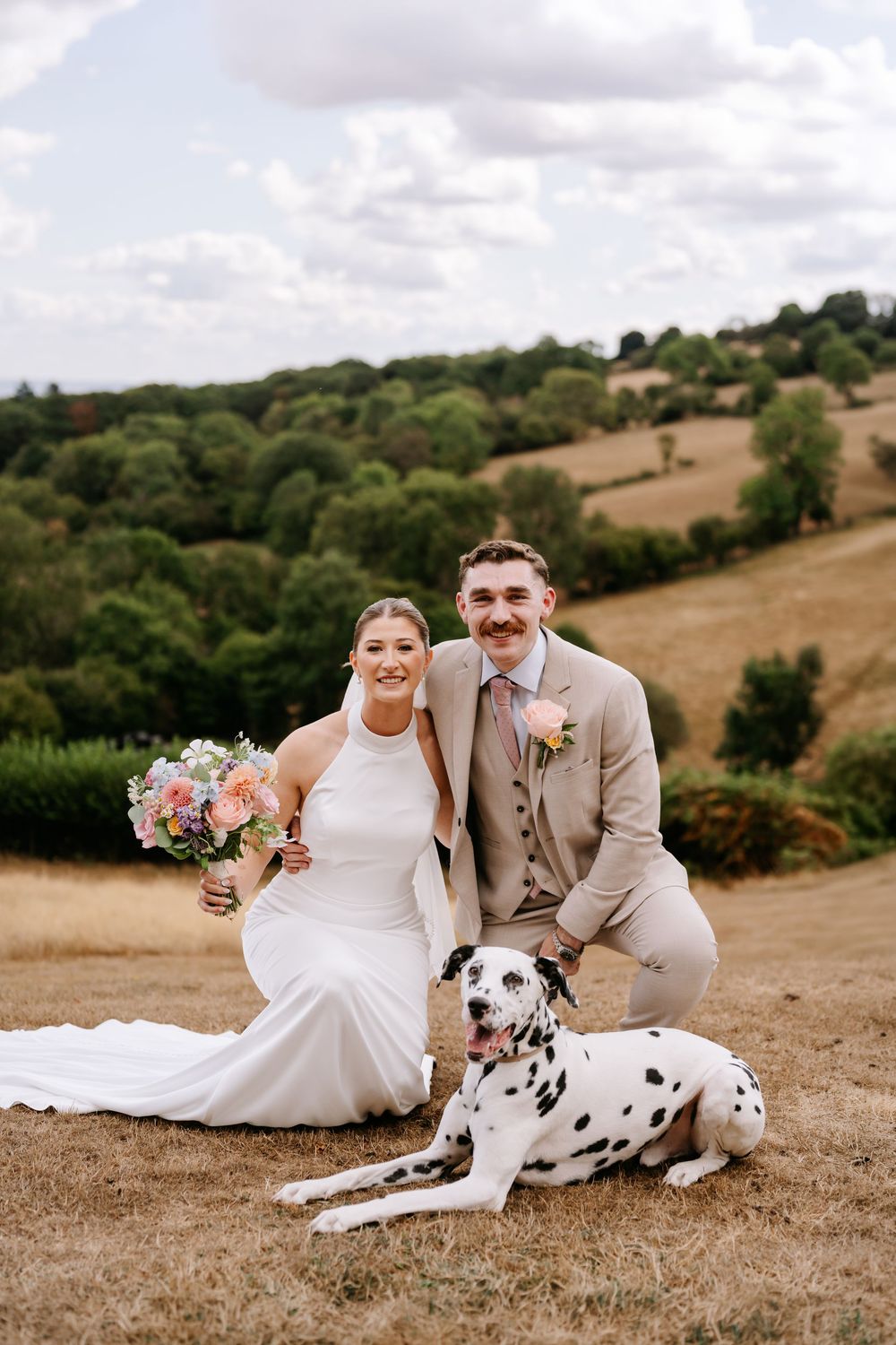 Bride and groom posing with a Dalmatian dog on a sunny hillside, bride holding a pastel bouquet in a white gown.