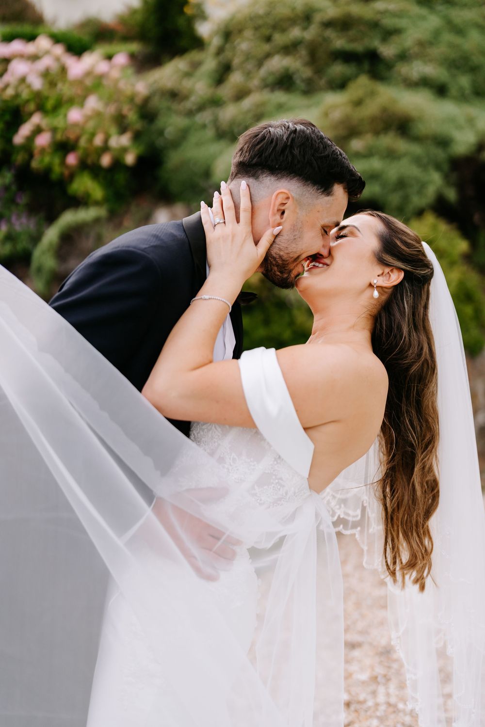 Bride and groom sharing a romantic kiss outdoors, with her veil flowing in the breeze and lush greenery behind them.