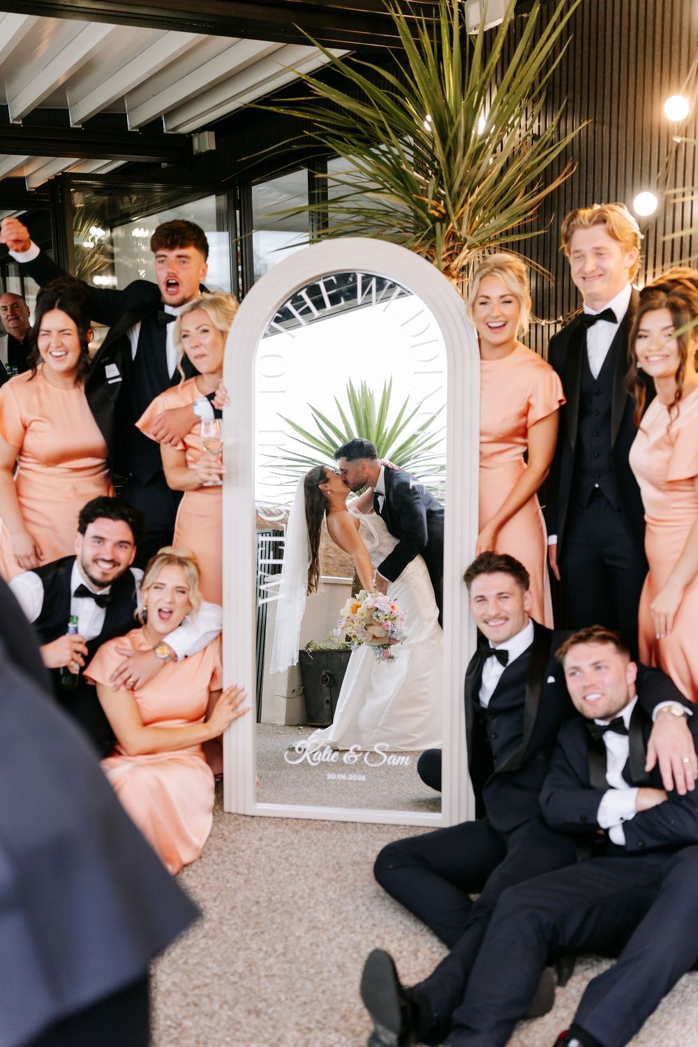 Wedding party surrounds a mirror reflecting the bride and groom kissing, bridesmaids in peach dresses and groomsmen in black tuxedos.