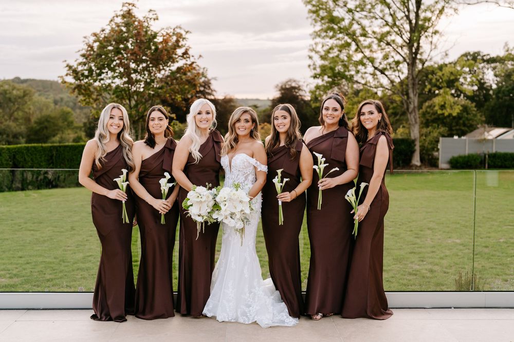 Bride in white lace gown poses with six bridesmaids in chocolate brown dresses holding white bouquets outdoors.