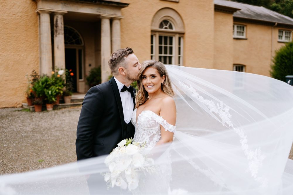 Bride and groom share a romantic kiss outside an elegant manor, her lace veil flowing beautifully in the breeze.