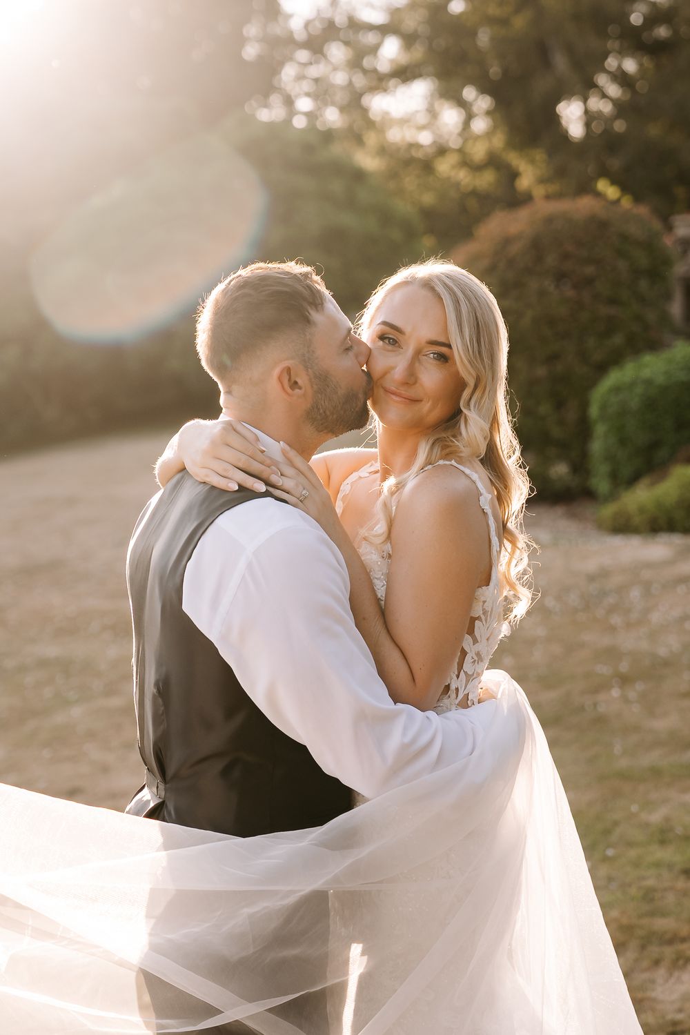 A bride and groom share a romantic embrace outdoors during golden hour, her veil flowing softly in the breeze.