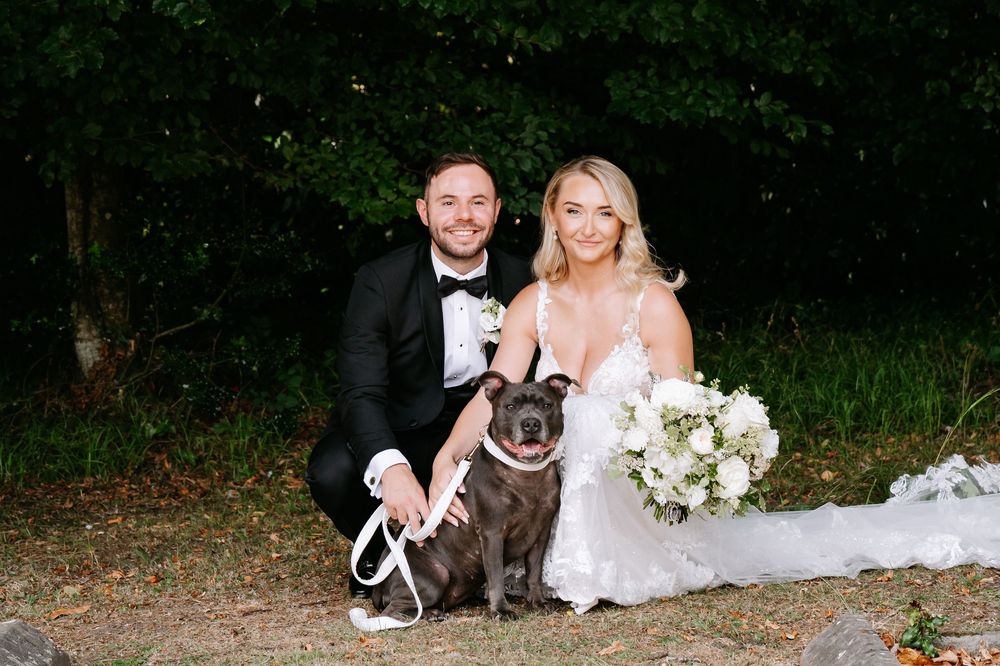 Bride and groom in wedding attire kneel beside their French Bulldog outdoors, bride holding white bouquet.