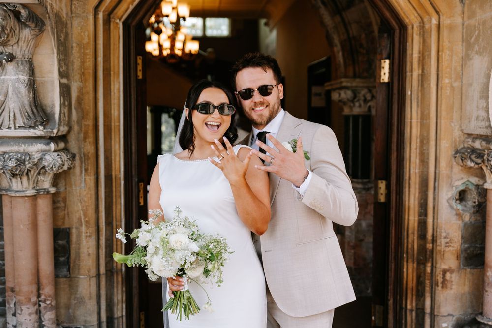 Bride and groom in sunglasses showing wedding rings outside ornate venue door, bride holding white floral bouquet.