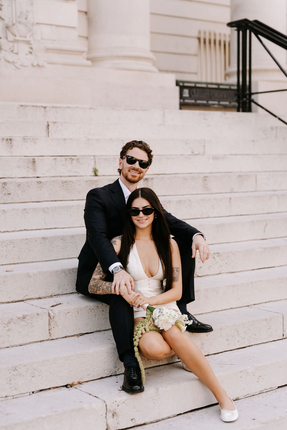 Couple in sunglasses sitting on marble steps, man in black suit, woman in white mini dress holding bouquet.
