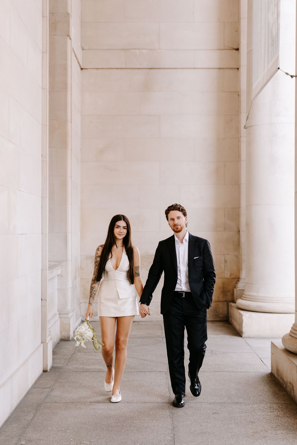 Couple in wedding attire walking hand-in-hand past stone columns, bride in white mini dress holding bouquet.