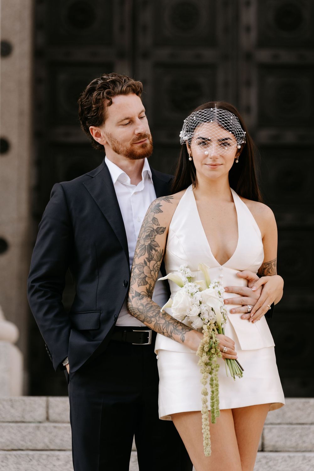 Tattooed bride in short white halter dress with birdcage veil holds bouquet beside groom in dark suit.