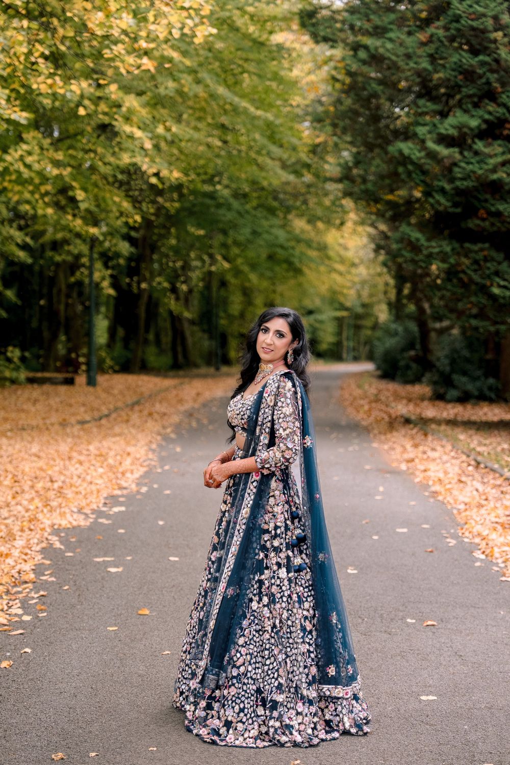Woman in embroidered teal lehenga poses on autumn tree-lined path, showcasing traditional South Asian bridal fashion.
