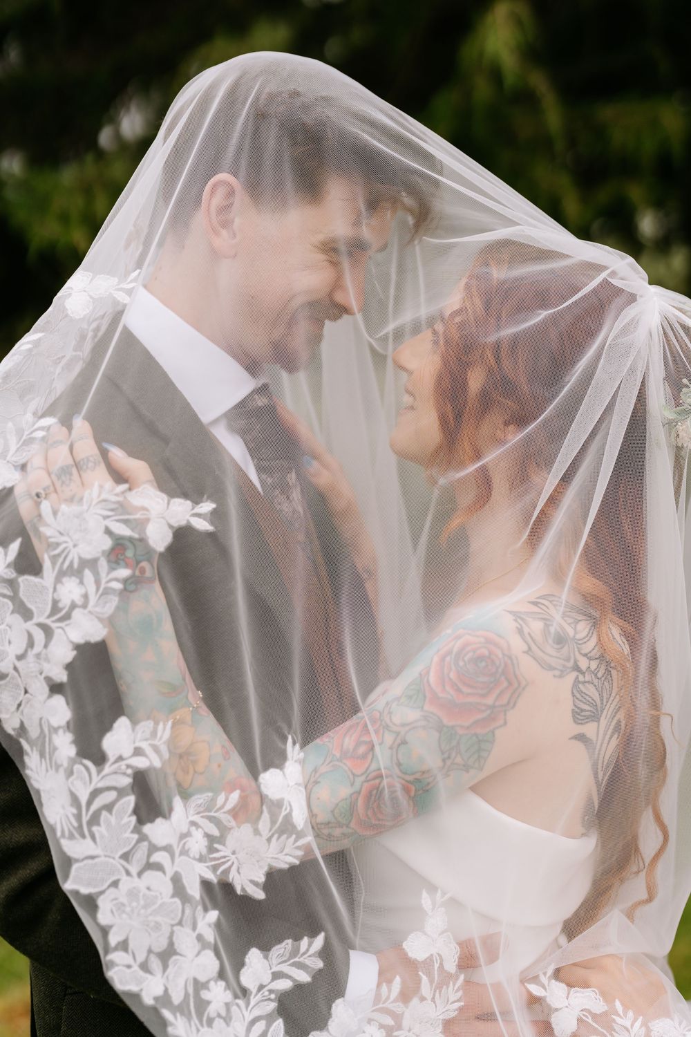 Bride and groom share romantic moment under lace wedding veil, groom in gray suit, bride with floral tattoos.