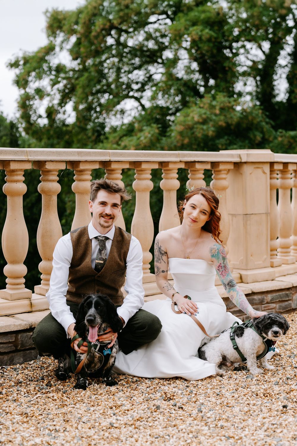 Smiling newlyweds in wedding attire sit with two dogs by stone balustrade in outdoor garden setting.