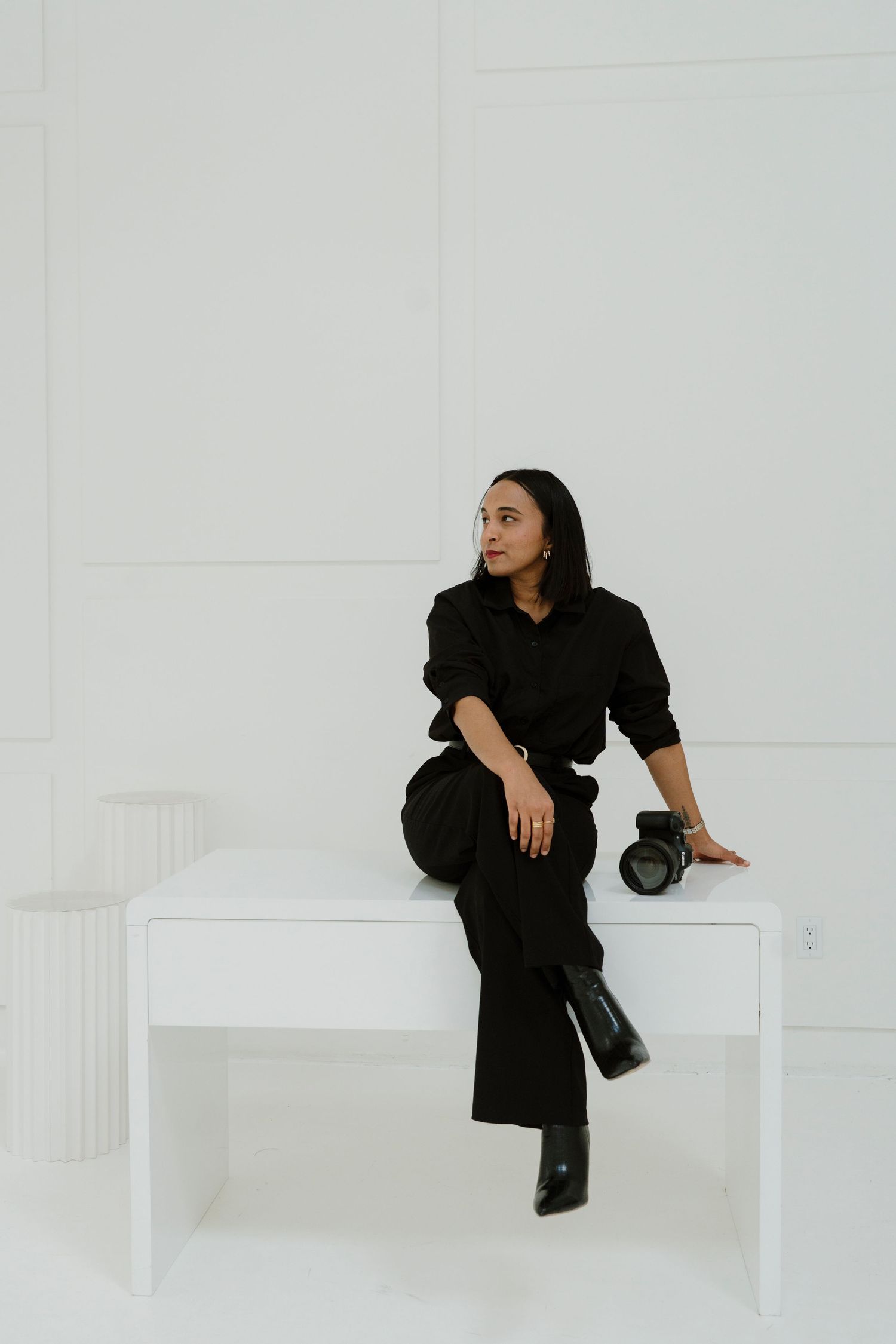 A person in all black attire sits on a white bench in a minimalist white studio setting with a camera nearby.