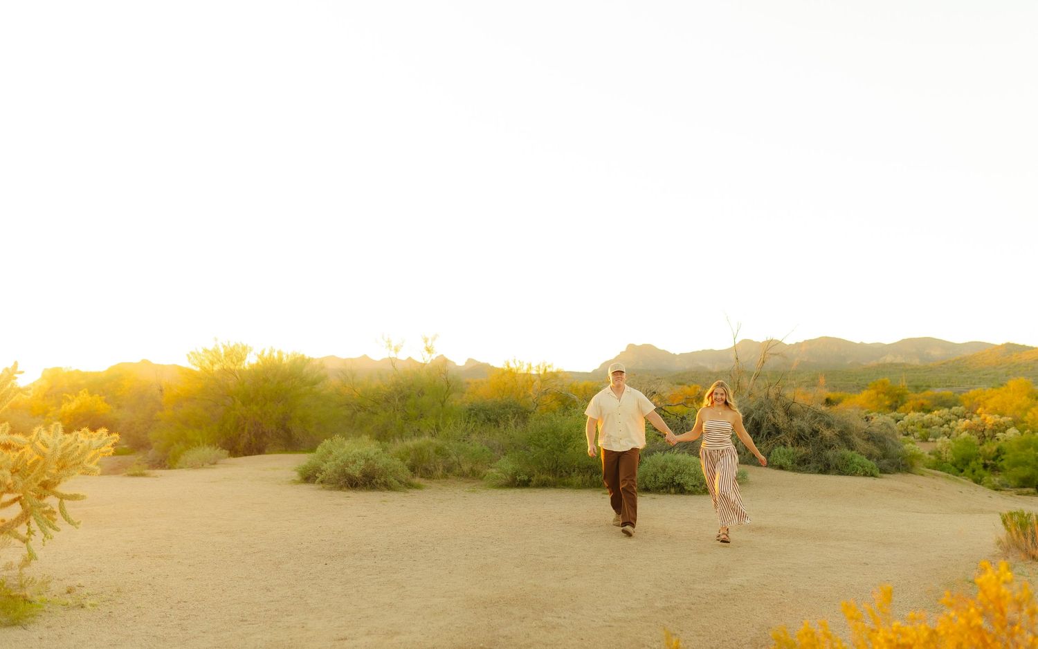 A newly married couple embrace each other in front of a lake, with their baby on his hip.