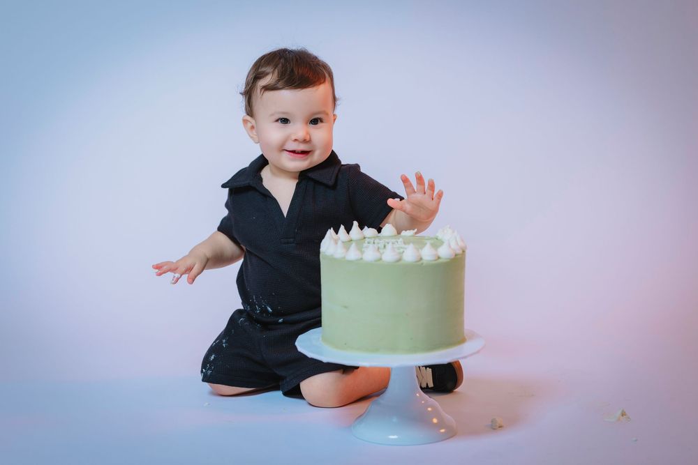 Cute baby in black outfit smiling beside green frosted cake on white cake stand in studio setting.