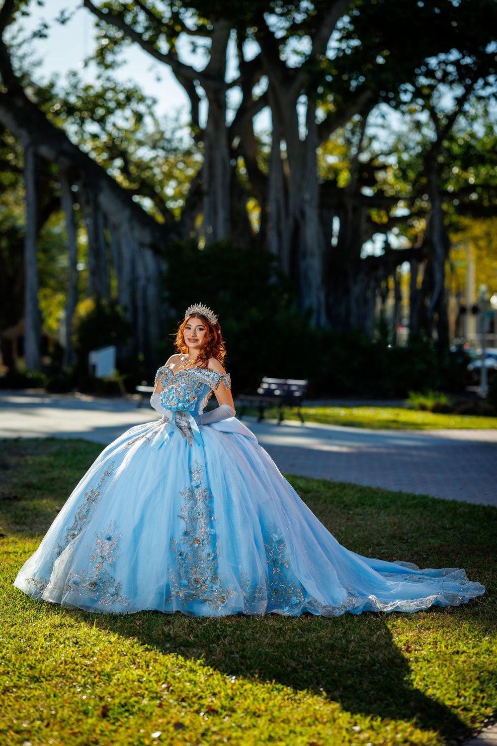 Young woman in light blue ball gown with floral embroidery and crown poses in sunlit park.