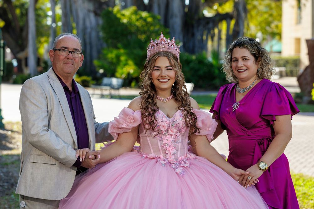 Girl in pink quinceañera gown and crown poses with parents outdoors in park setting.