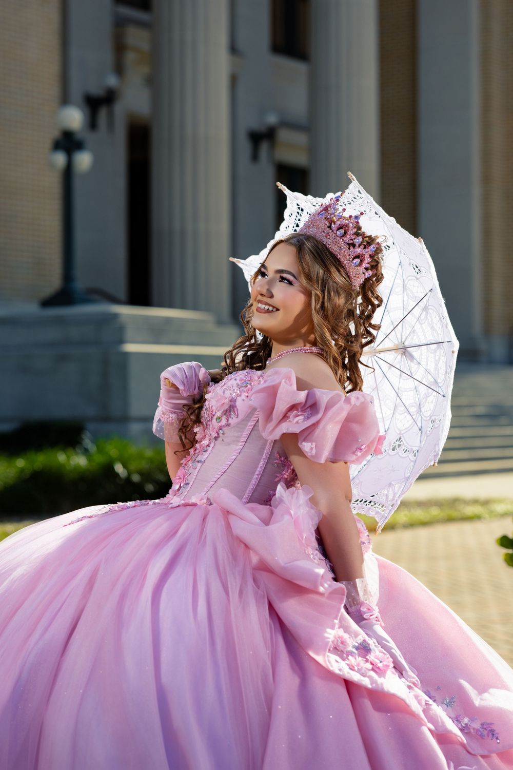 Young woman in pink ball gown with lace umbrella and crown poses outdoors near classical building.