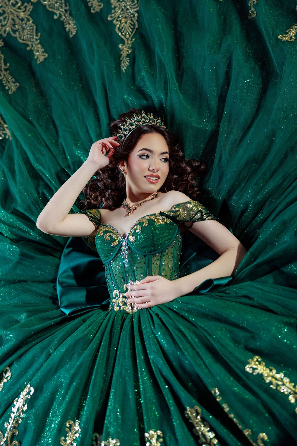 Young woman in emerald green and gold ball gown with crown lies surrounded by flowing skirt.