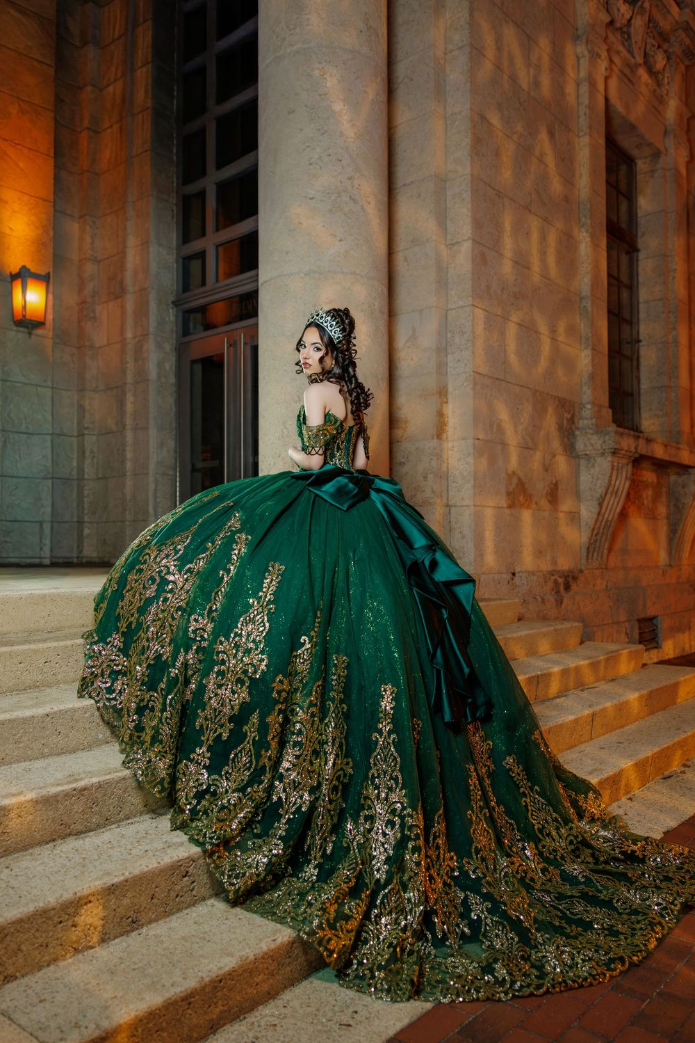 Young woman in green and gold quinceañera gown poses on steps of ornate building at night.