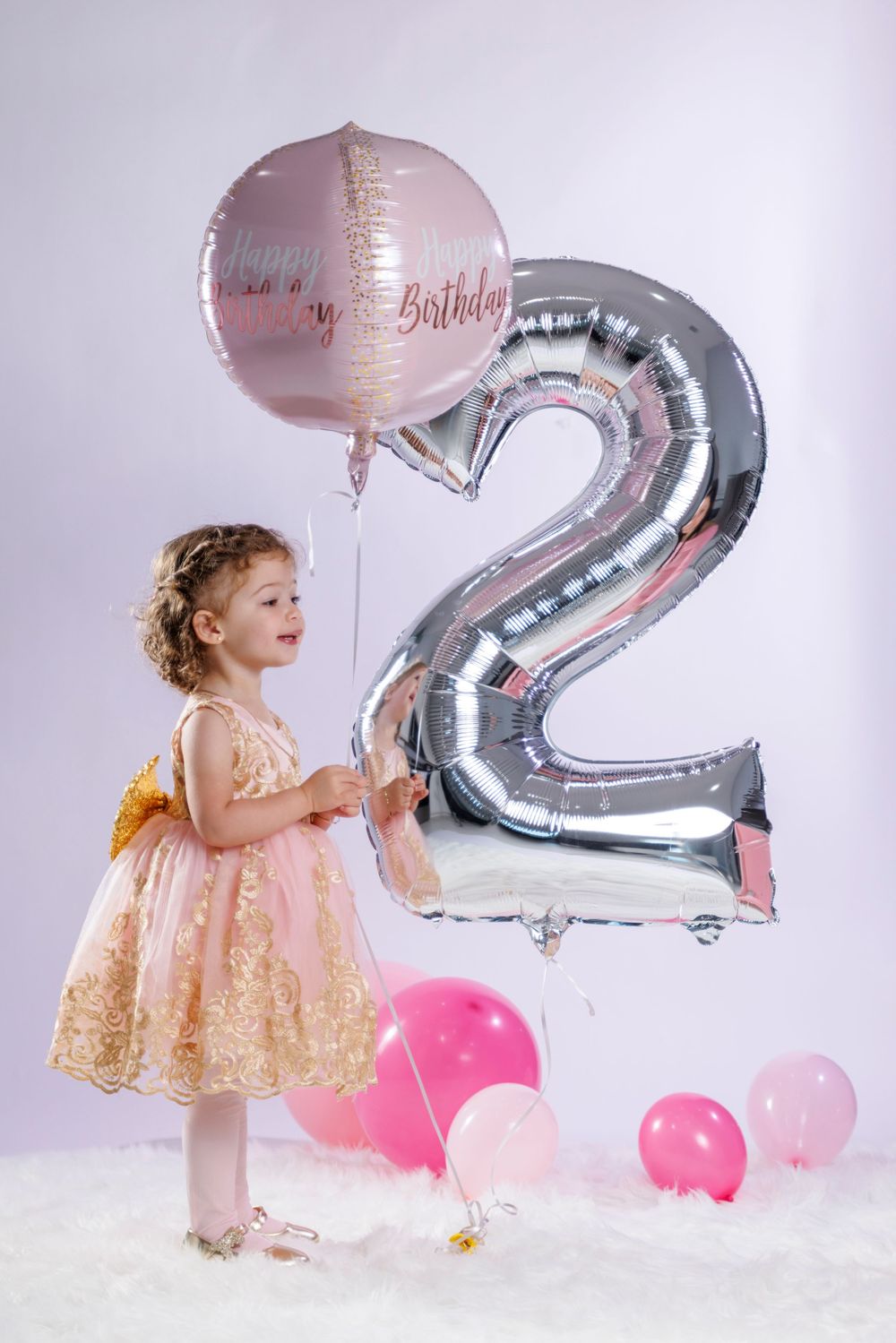 Little girl in pink gold dress holding Happy Birthday balloon next to large number 2 silver balloon.