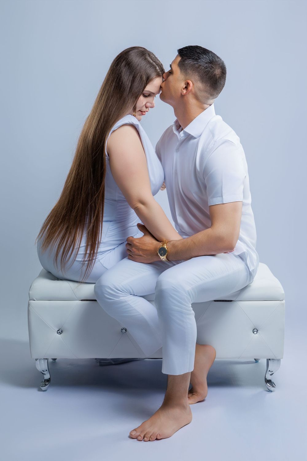 Romantic couple in white outfits sitting close together sharing an intimate moment on white bench.