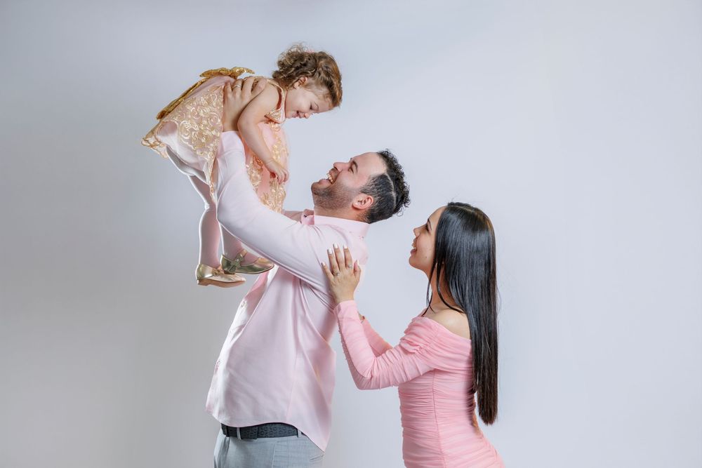 Joyful family portrait with father lifting toddler daughter while mother watches lovingly in pink outfits.