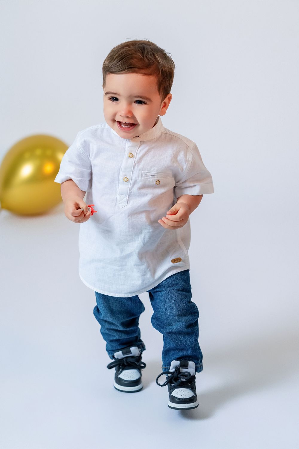 Happy baby boy in white shirt and jeans standing and smiling with gold balloon in white studio background.