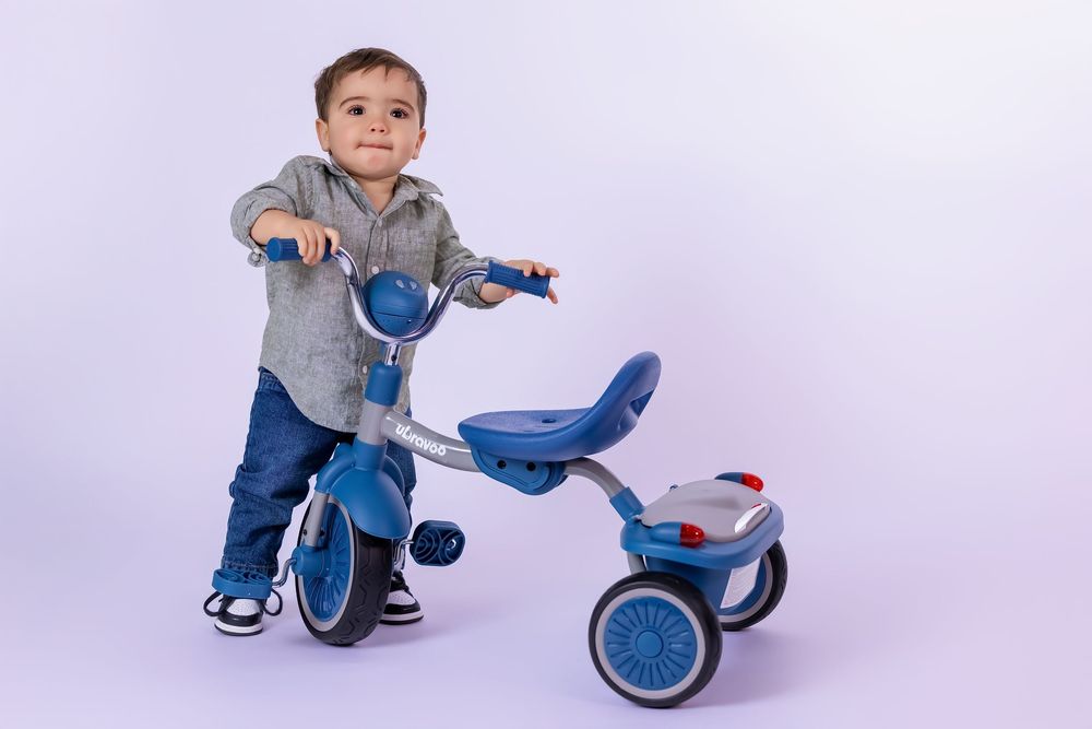 Toddler boy riding blue tricycle, wearing gray shirt and jeans, smiling at camera on white background.