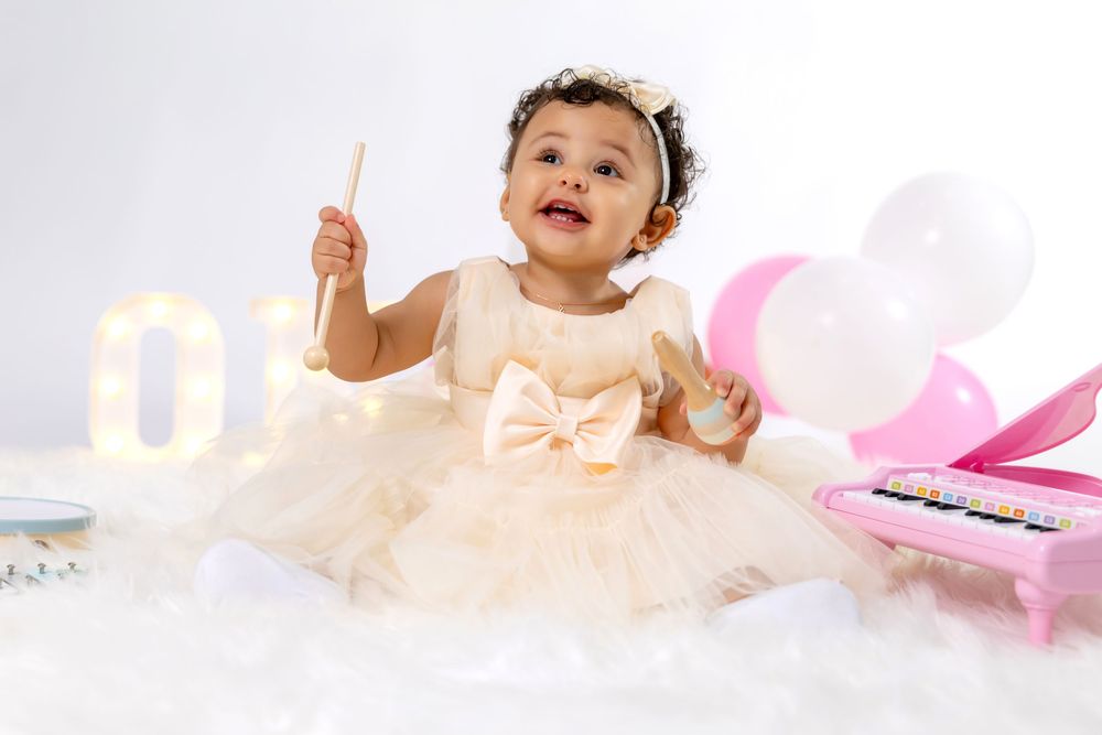 Adorable baby girl in cream tutu dress with bow sitting among balloons and toys celebrating first birthday.