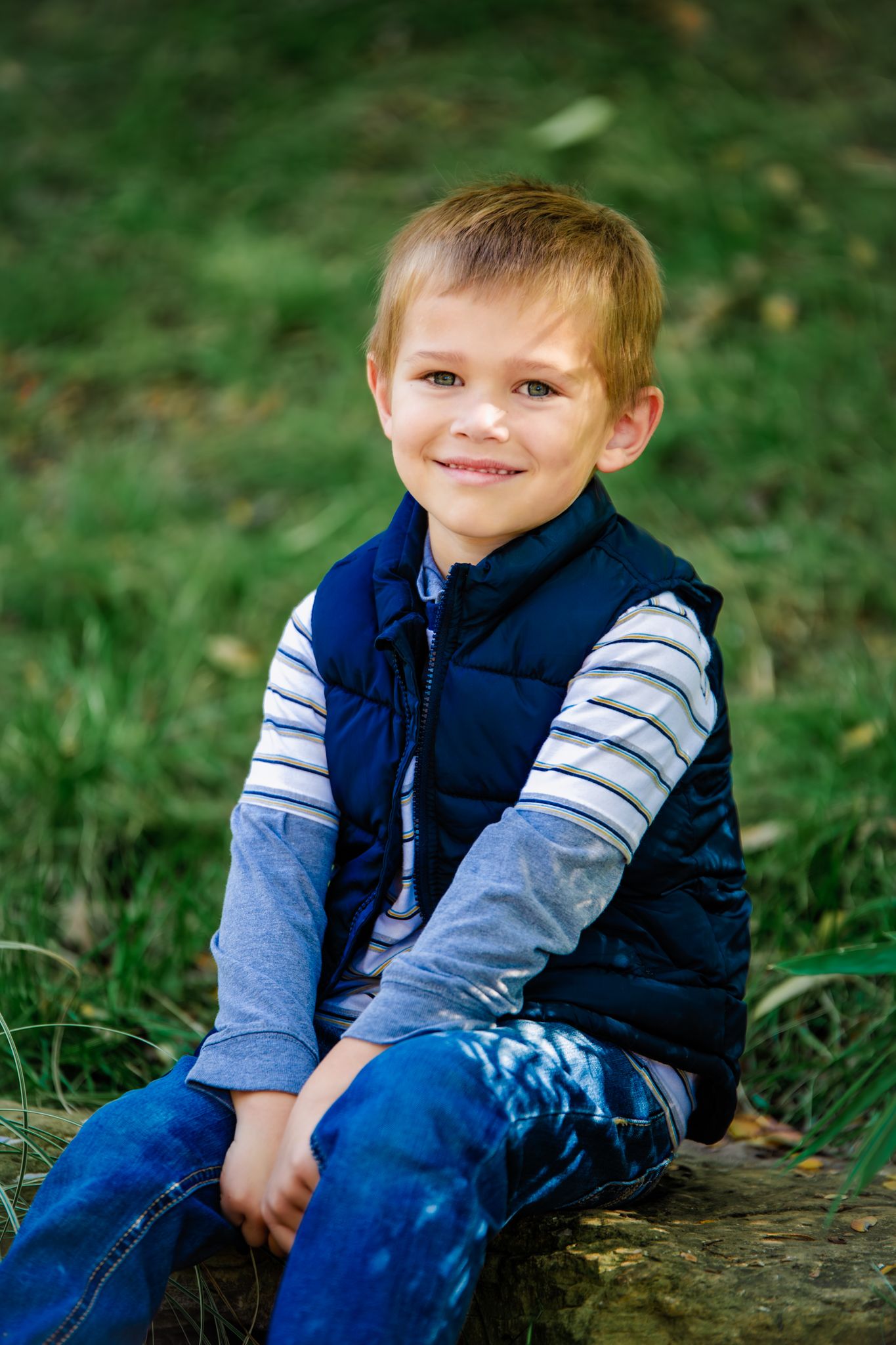 A child in a navy vest and striped sleeves sits casually on grass during an outdoor portrait session.