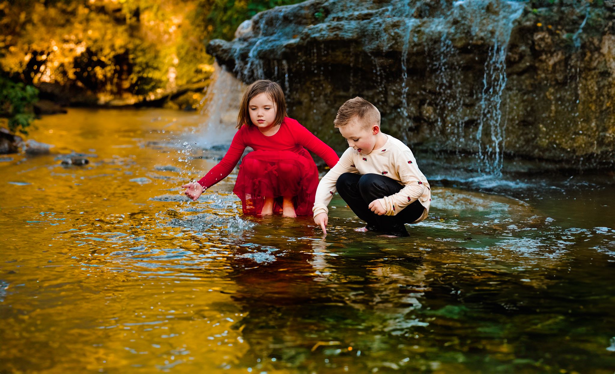 Children play and splash in shallow golden-colored water near rocks on a sunny day.