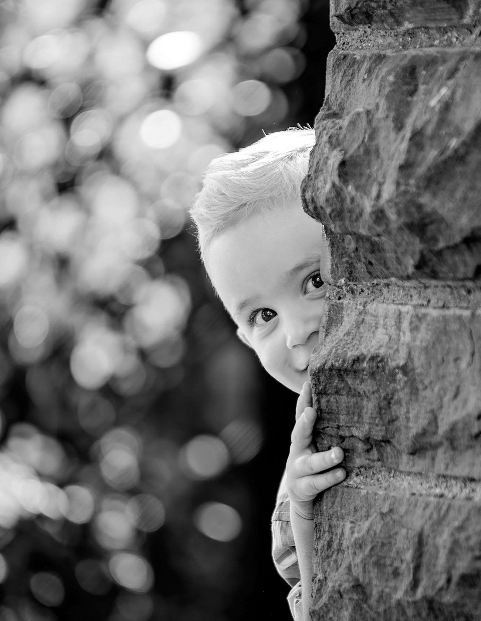 Black and white artistic portrait series showing a playful peek around a textured stone wall with soft bokeh background.