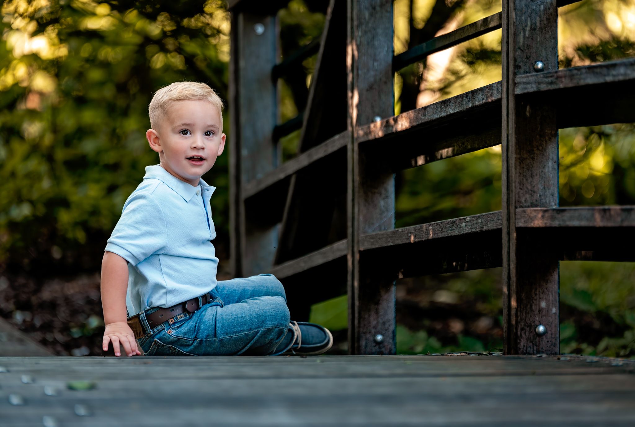 A young child in blue shirt and jeans sits on a wooden bridge during golden hour lighting.