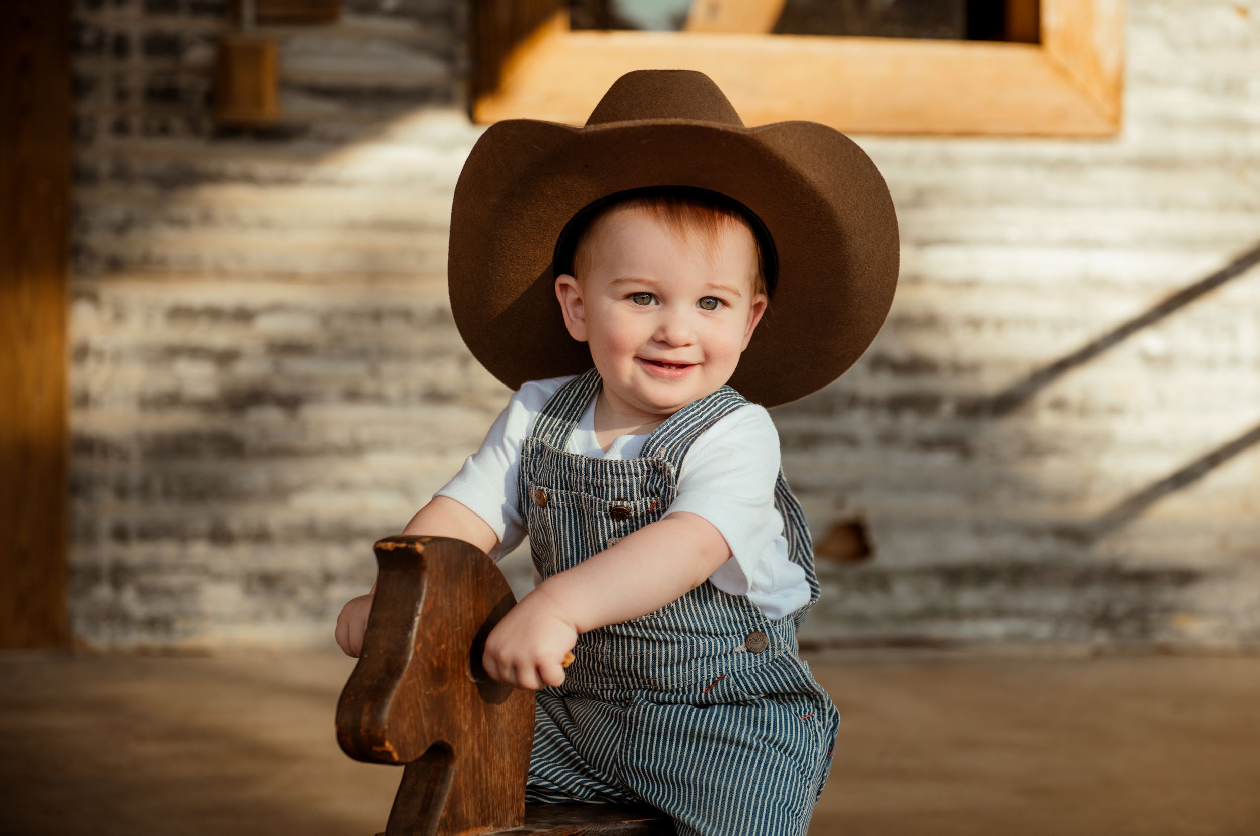 Small child wearing denim overalls and brown cowboy hat sits on wooden rocking horse in rustic setting.