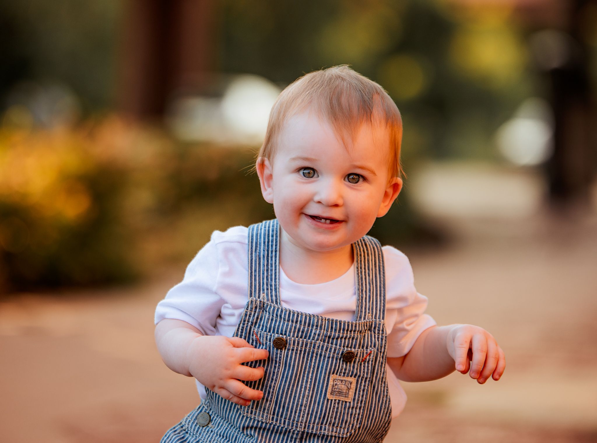 A small child wearing denim overalls sits and plays outdoors during autumn with fallen leaves in the background.