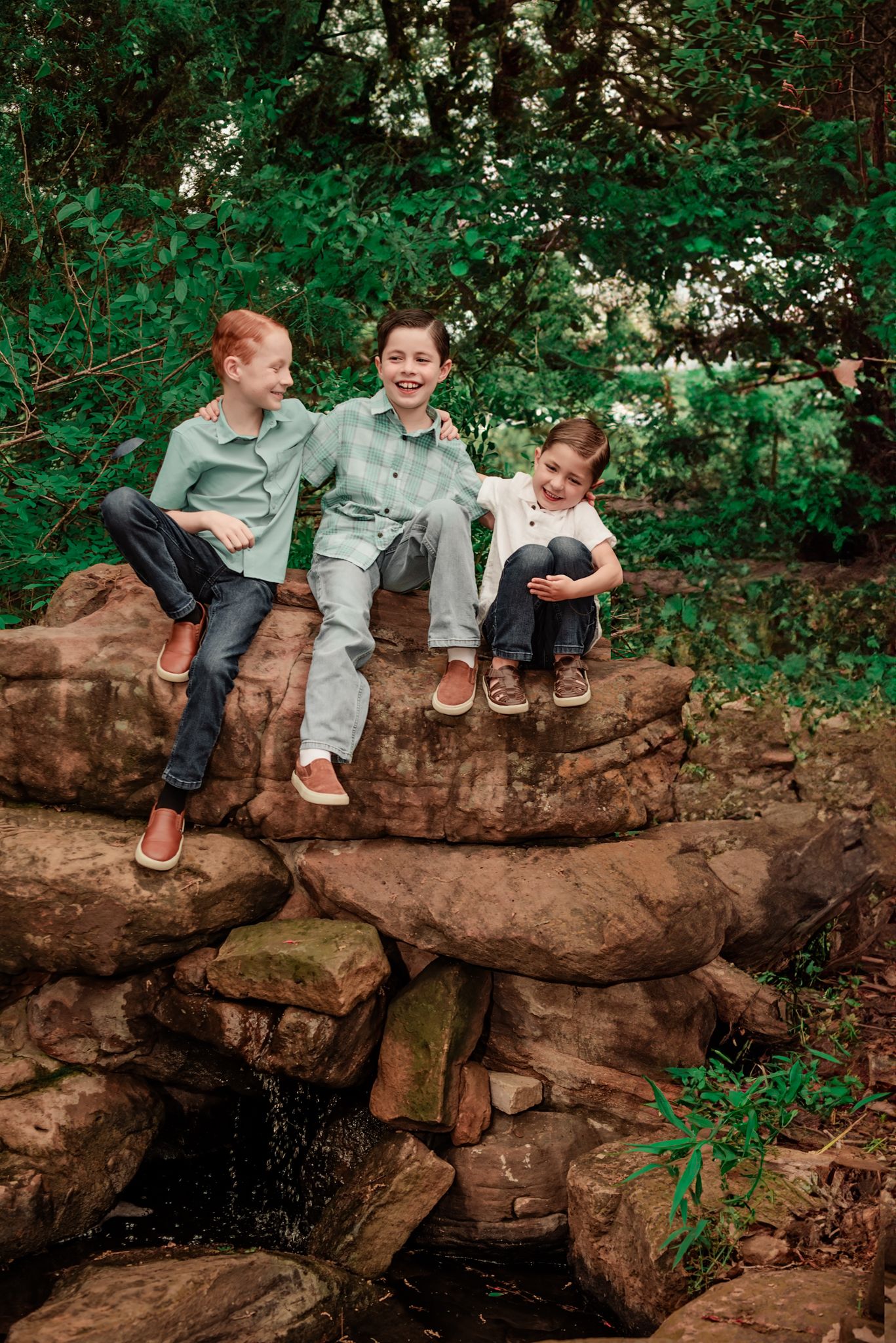 Two children in casual clothing sit and laugh together on a large fallen tree trunk surrounded by lush green foliage.