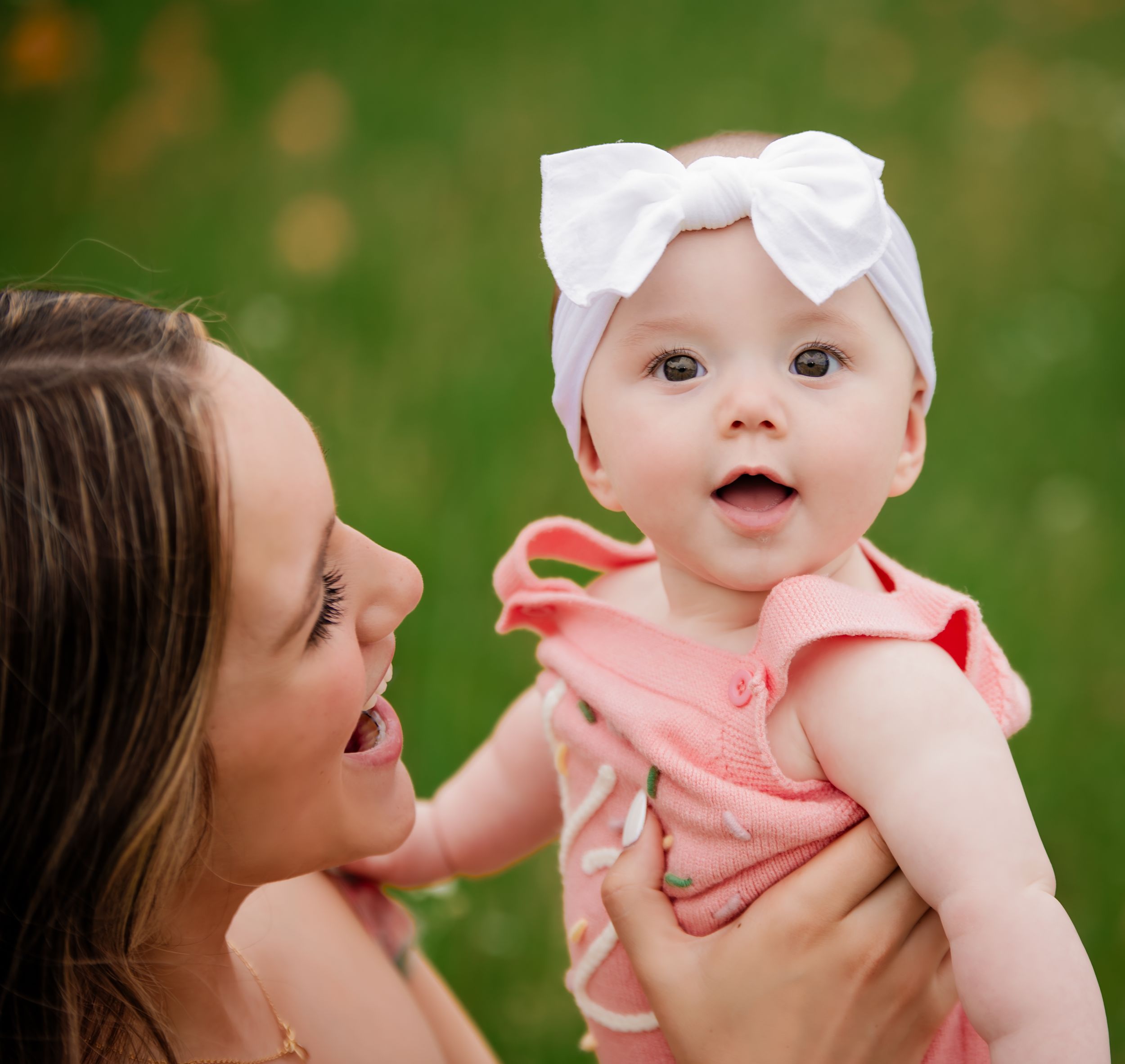 Baby wearing a white bow headband and peach dress smiles joyfully while being held up outdoors on a sunny day.