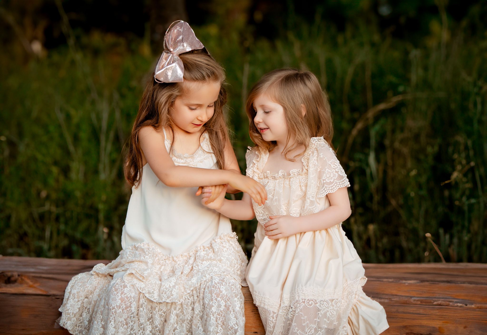 Two young children in white lace dresses sit together on a wooden bench in a garden setting.