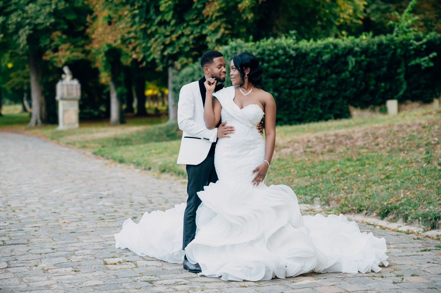A bride in a white gown and groom in black and white tuxedo hold hands while walking outdoors in a park setting.