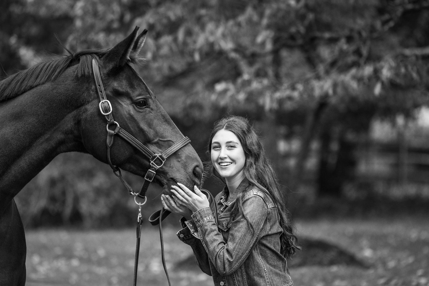 Black and white photo of a smiling young woman with long hair gently holding a horse's face outdoors.