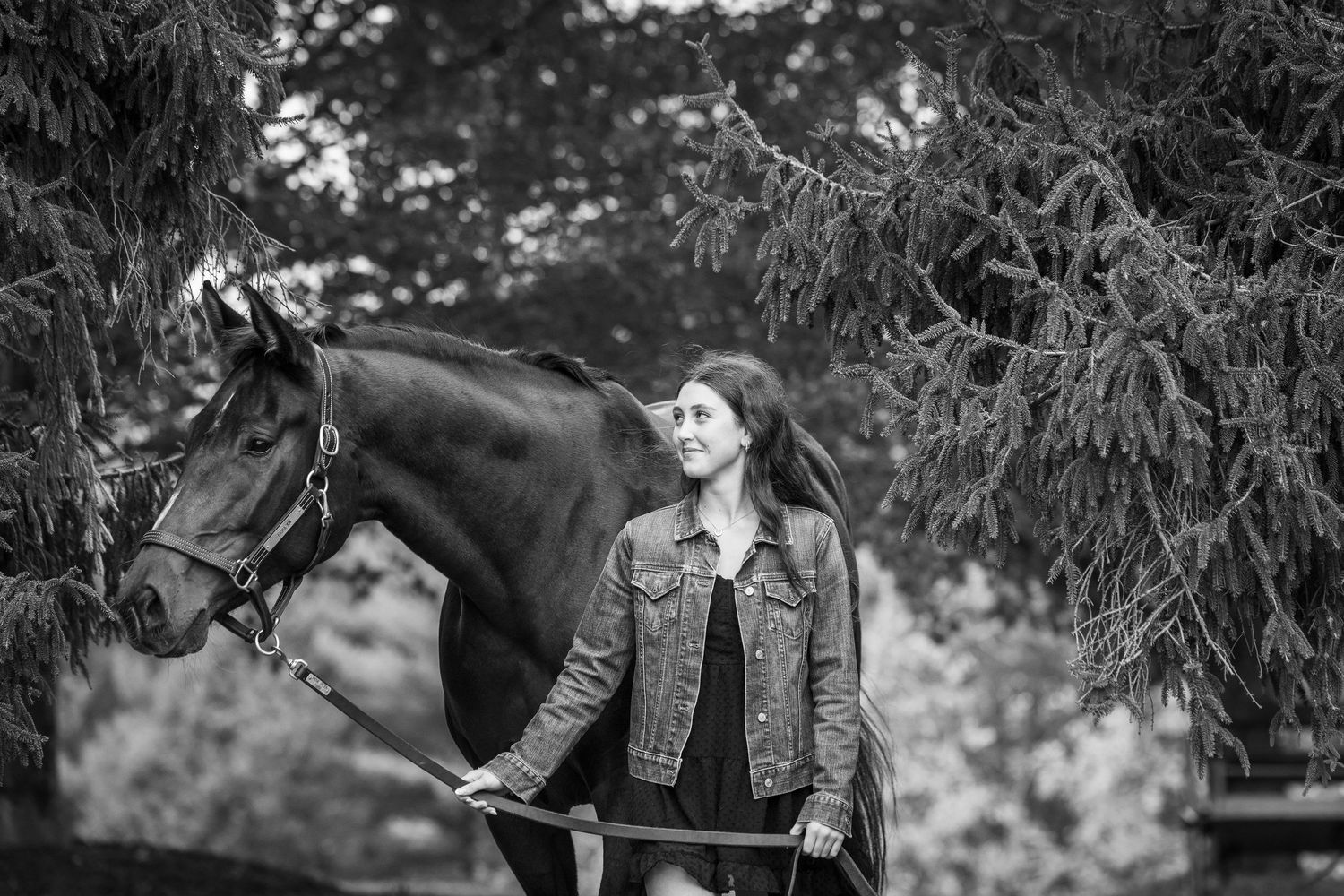 Black and white photo of a smiling young woman in a denim jacket standing beside a dark horse under pine trees.