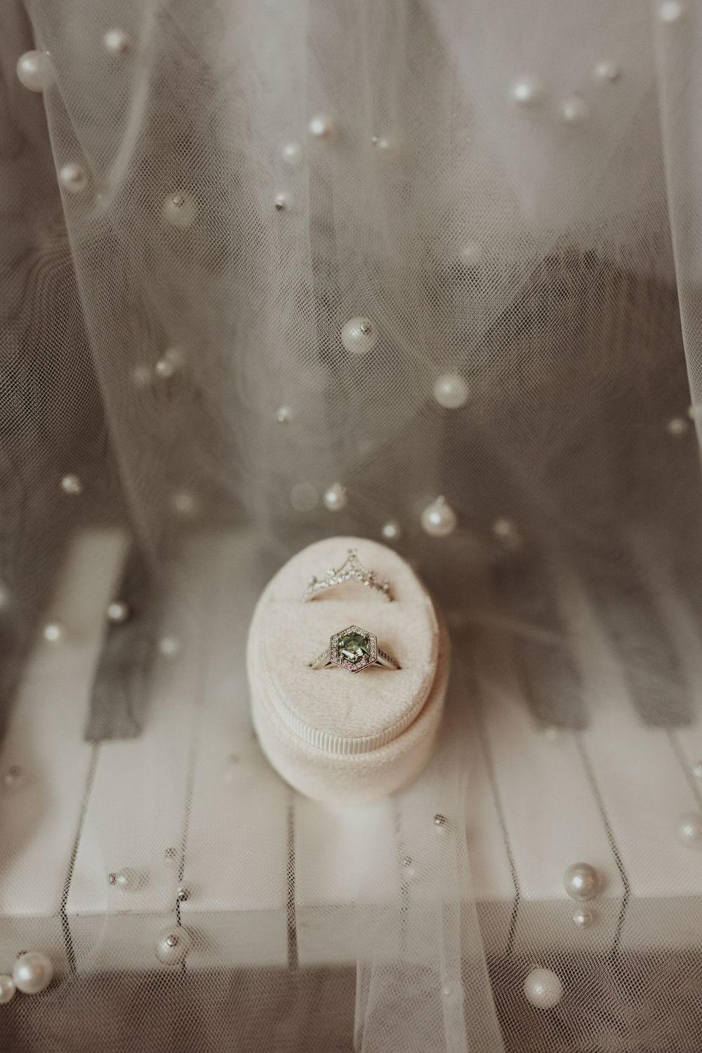 Wedding ring rests on a white cushion surrounded by delicate tulle and pearls in a dreamy monochrome setting.