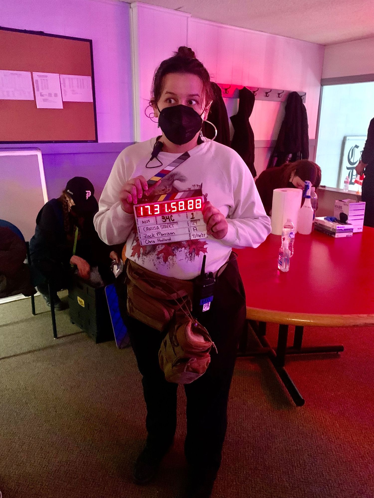 Crew member wearing a white hoodie and mask in a pink-lit production room.