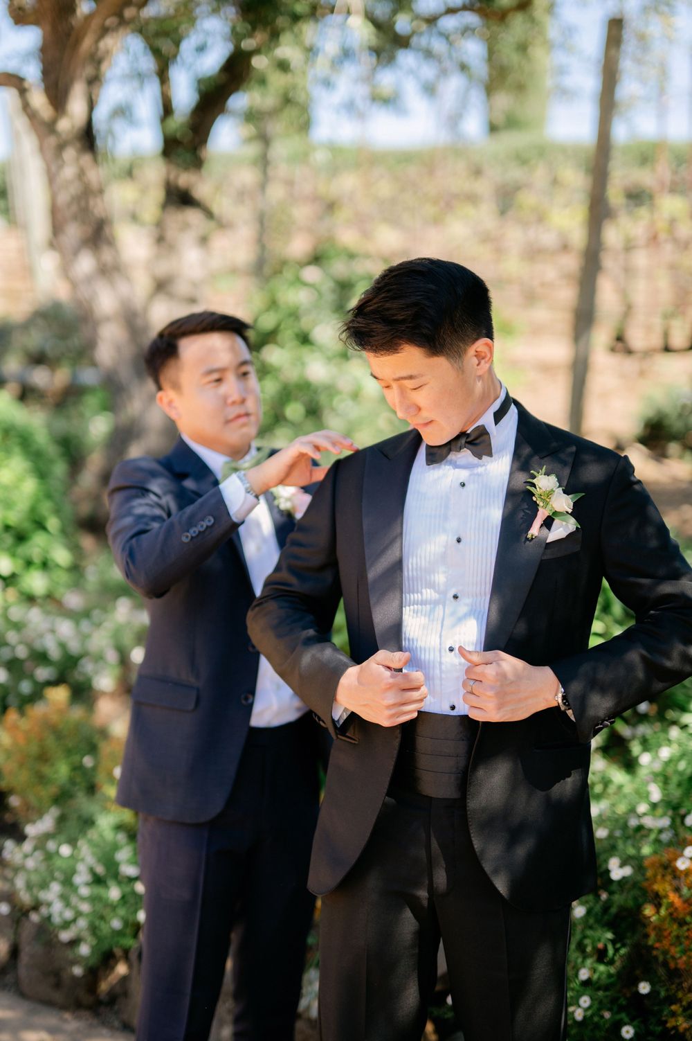 Final adjustments being made to the groom's formal attire in an outdoor garden setting.