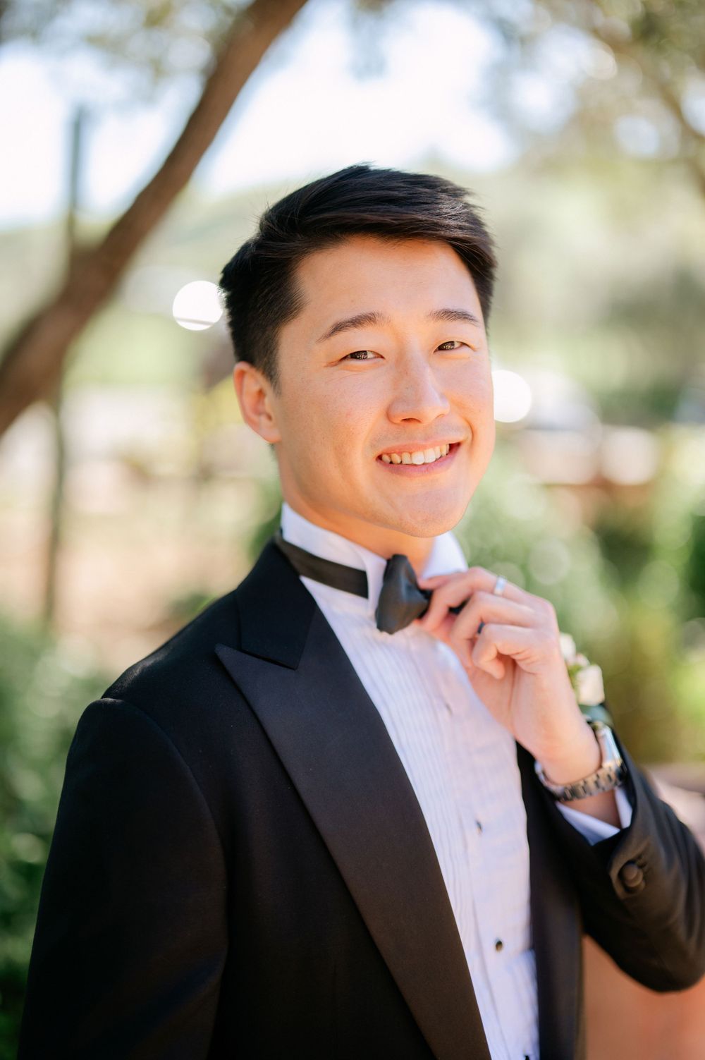 Groom in classic black tuxedo adjusting bow tie while smiling outdoors.