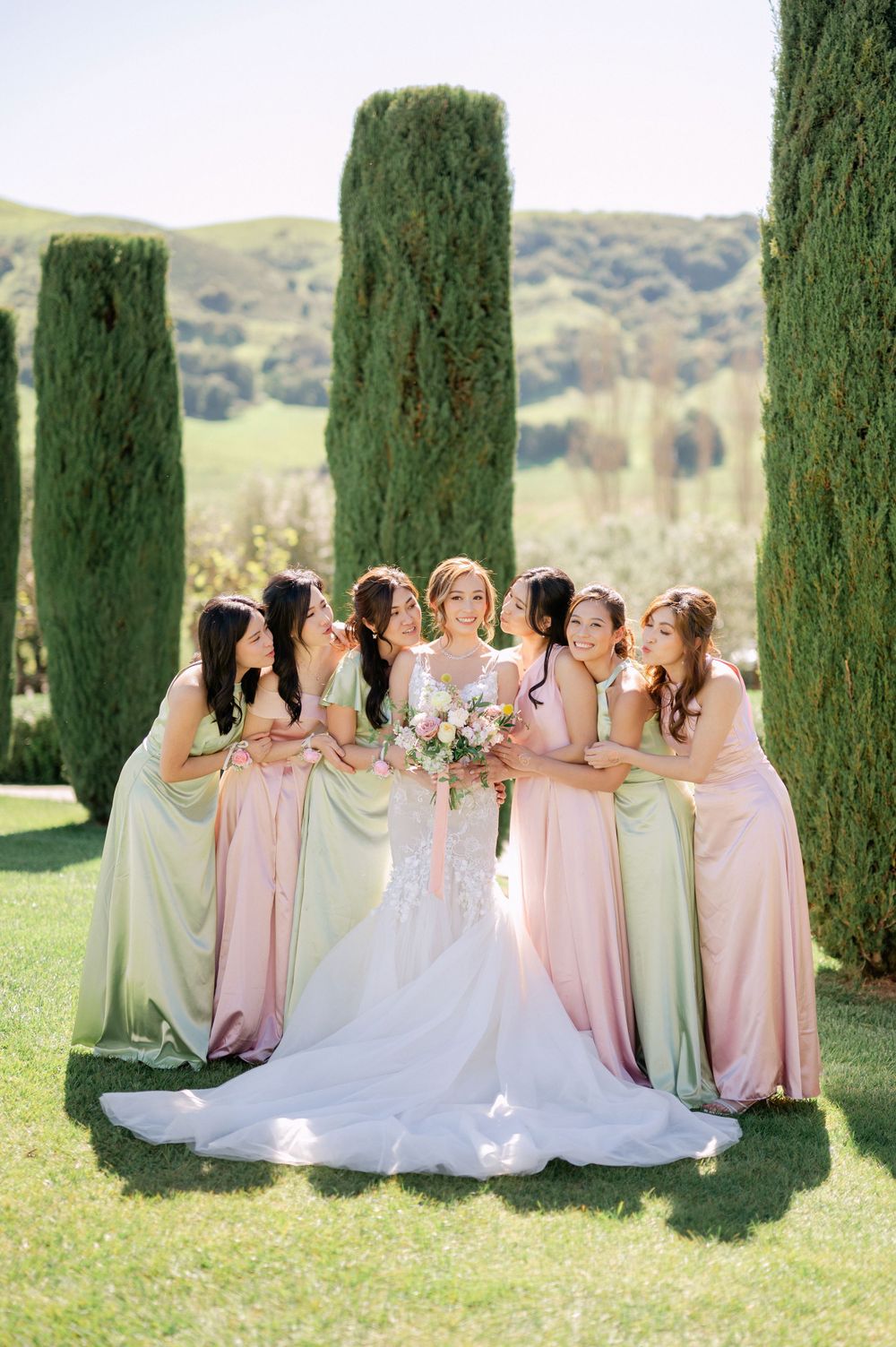Wedding party gathers intimately around bride in white gown framed by majestic cypress trees.