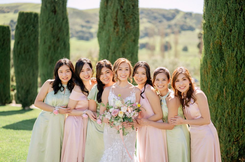 Bridesmaids in pastel pink and mint dresses pose with bride against towering cypress trees in elegant garden setting.